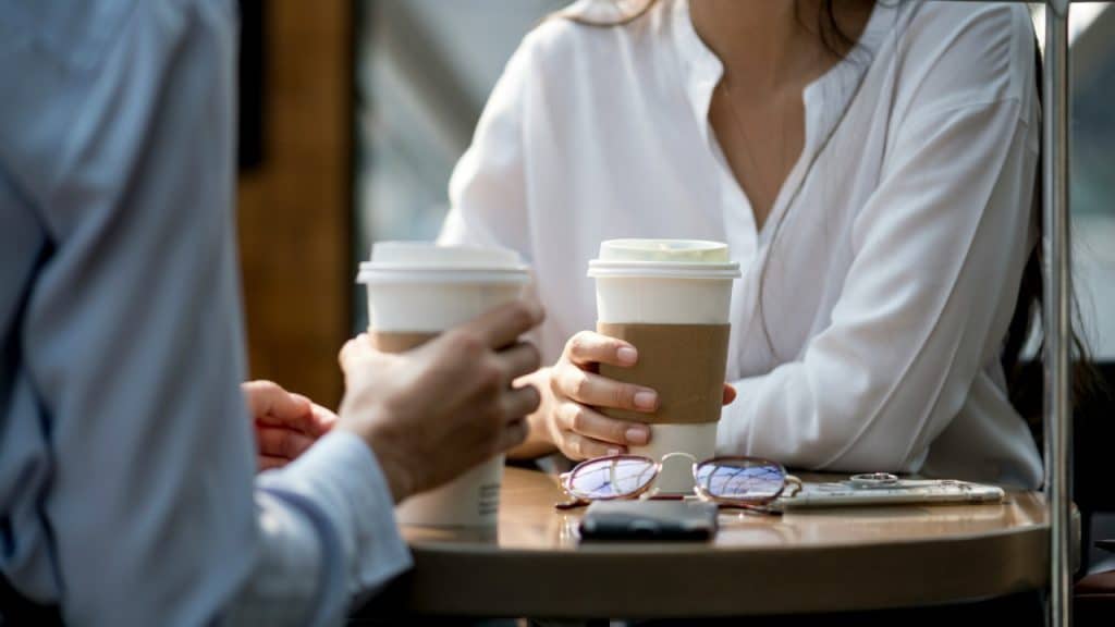Two people hold paper coffee cups while sitting at a small round table.