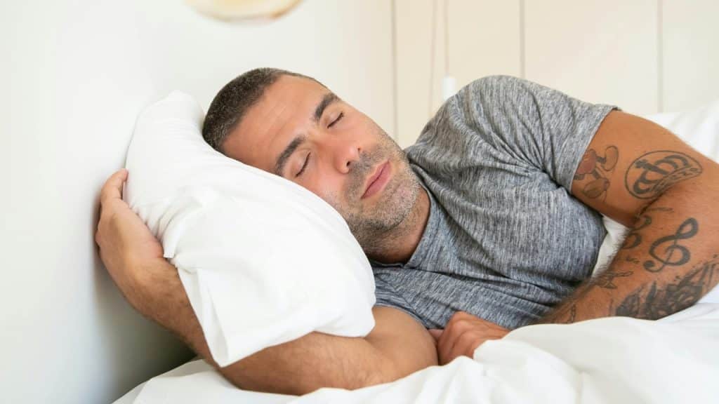 A man with short hair and arm tattoos sleeps on a white pillow and bedding.