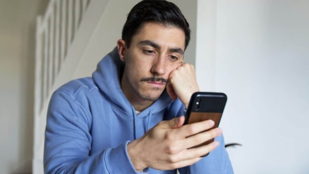 A man in a blue hoodie rests his chin on his hand while looking at a smartphone.