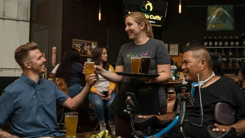 A waitress serving drinks to two men smiling at a bar.