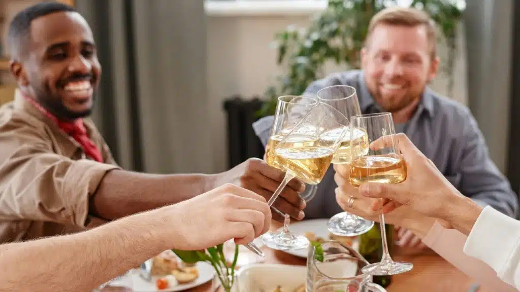 Several people around a table toast with glasses of white wine during a social gathering indoors.
