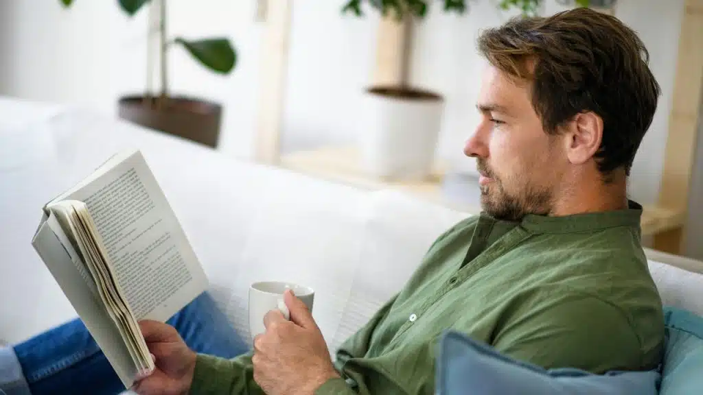 A man holds a mug and reads an open book while sitting on furniture.