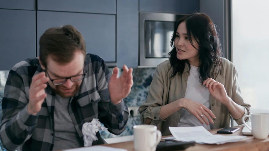 A man with glasses holds his head while a woman speaks at a table.