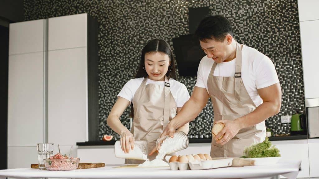 A woman and man in aprons prepare dough on a table in a kitchen.