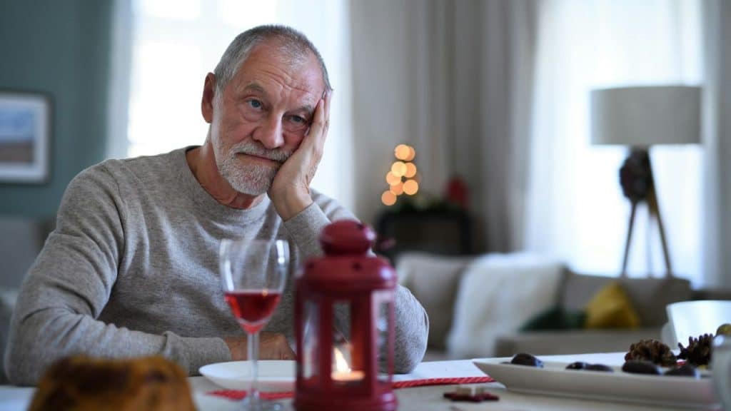 An older man sits at a table with a glass of wine, looking thoughtful.