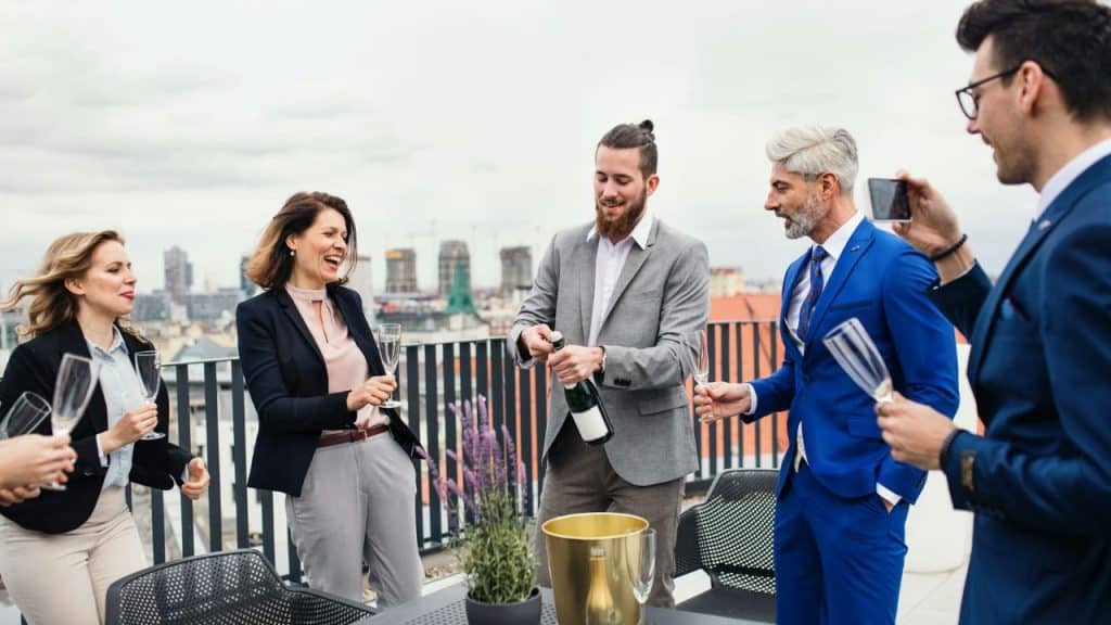 A group of people in formal attire stand on a balcony while opening a bottle.