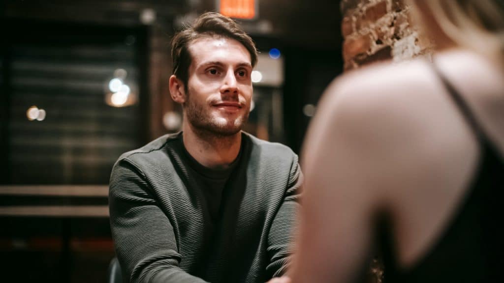 A man looking off-camera sits across from a woman in a dimly lit room.