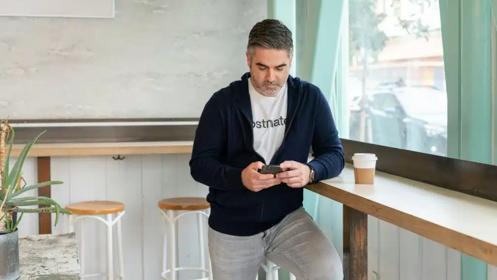 A man with graying hair leans against a counter, focused on his mobile phone screen.