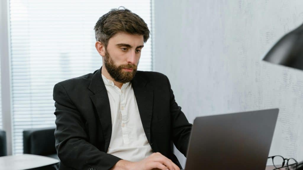 A bearded man in a black jacket and white shirt works on a gray laptop.