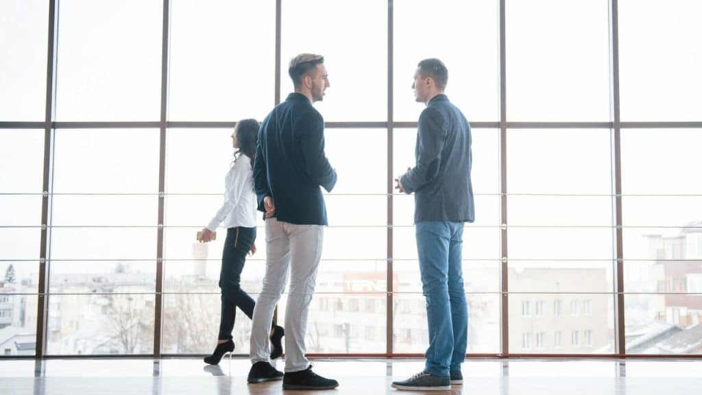 Two men talking in a hallway while a woman walks by in the background.