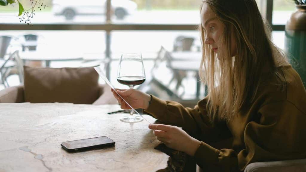 A woman reading a paper while sitting at a table with a glass of wine and a phone nearby.