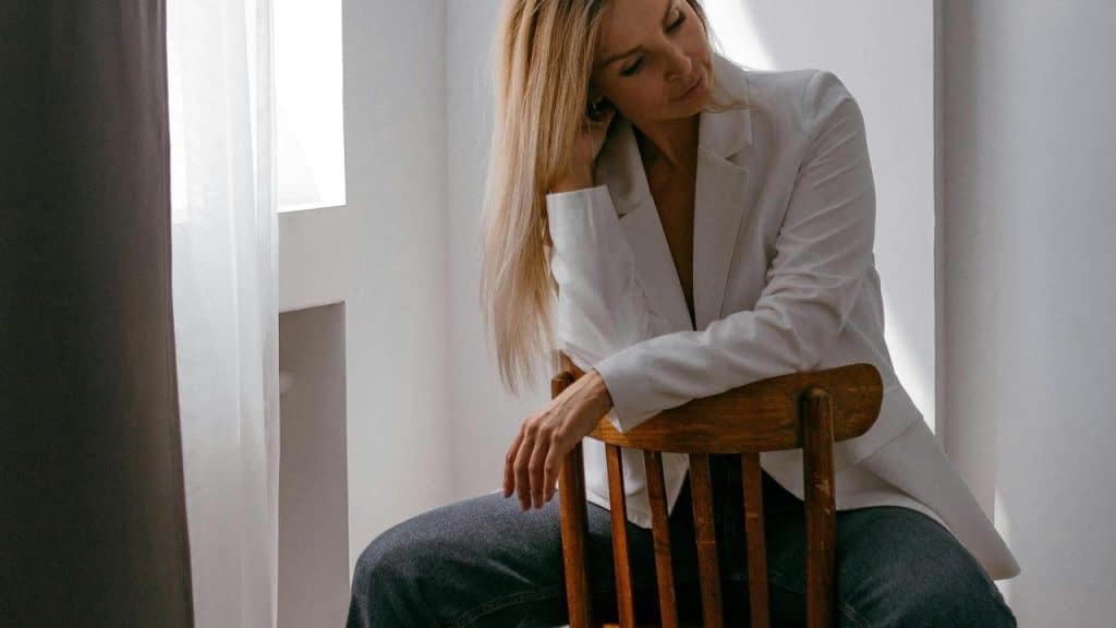 A woman in a white blazer and blue jeans sitting on a wooden chair.