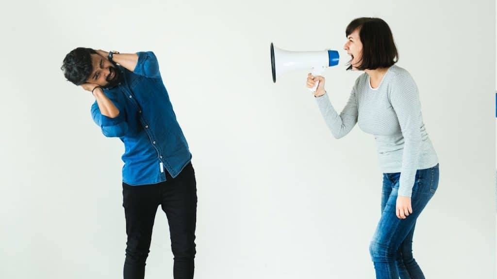 A man in a blue denim shirt covering his ears while a woman in a striped top shouts into a megaphone against a white background.