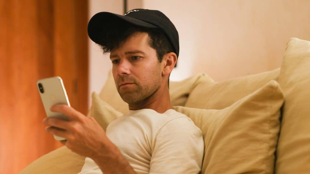 A man wearing a baseball cap sits on a sofa, looking at his smartphone.