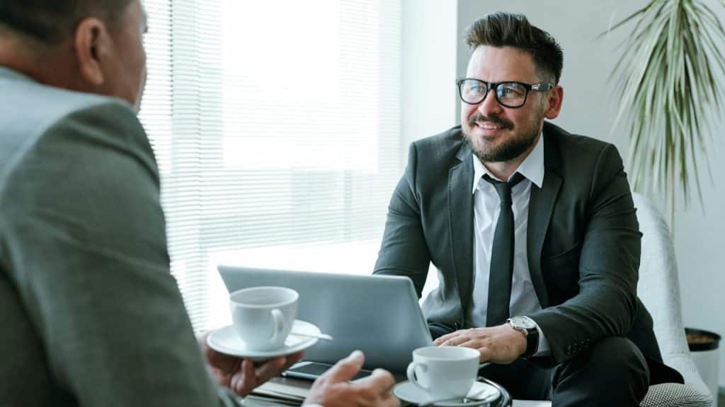 Two businessmen in suits sit together at a table with laptops and coffee.