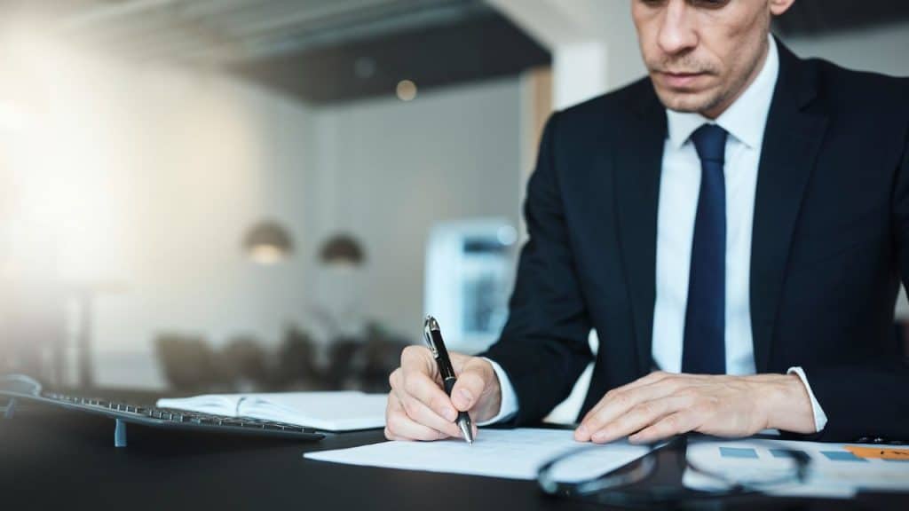A man in a suit and tie sits at a desk signing a document.