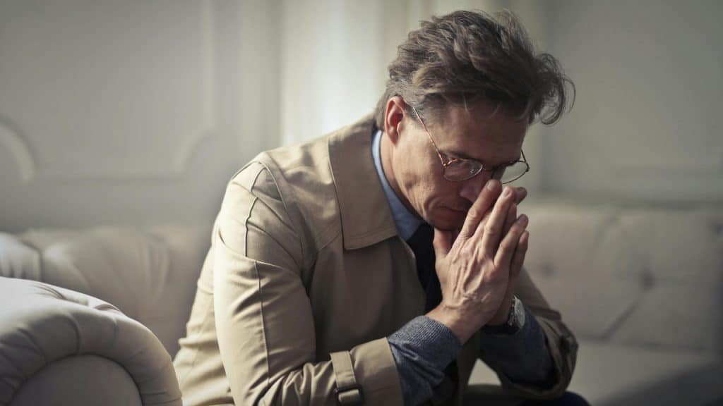 A man in a coat sits with his hands clasped, looking down, appearing distressed.
