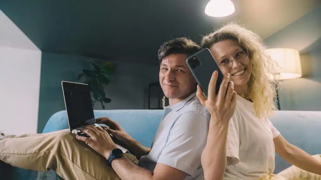 A couple sitting back-to-back on a couch using a laptop and smartphone.