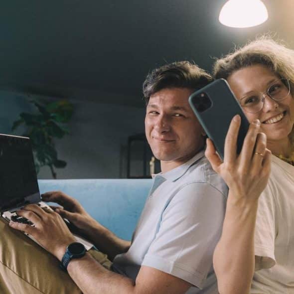 A couple sitting back-to-back on a couch using a laptop and smartphone.