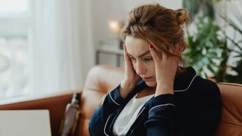 A woman sits on a couch with her hands on her head, looking distressed.