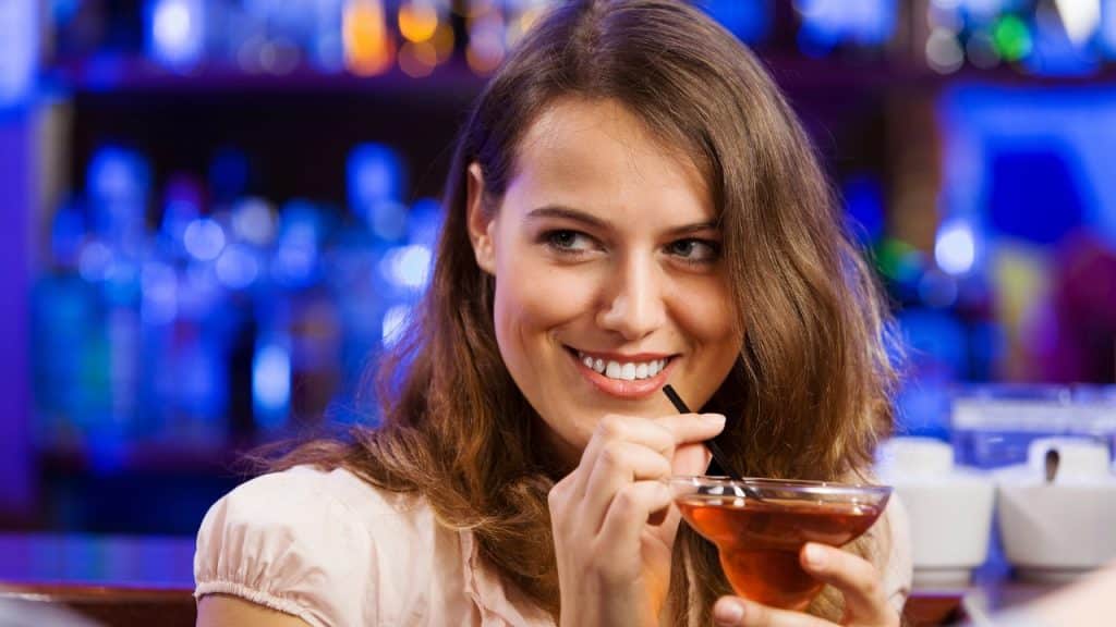 A woman with brown hair smiles while holding a glass with a straw in a bar.