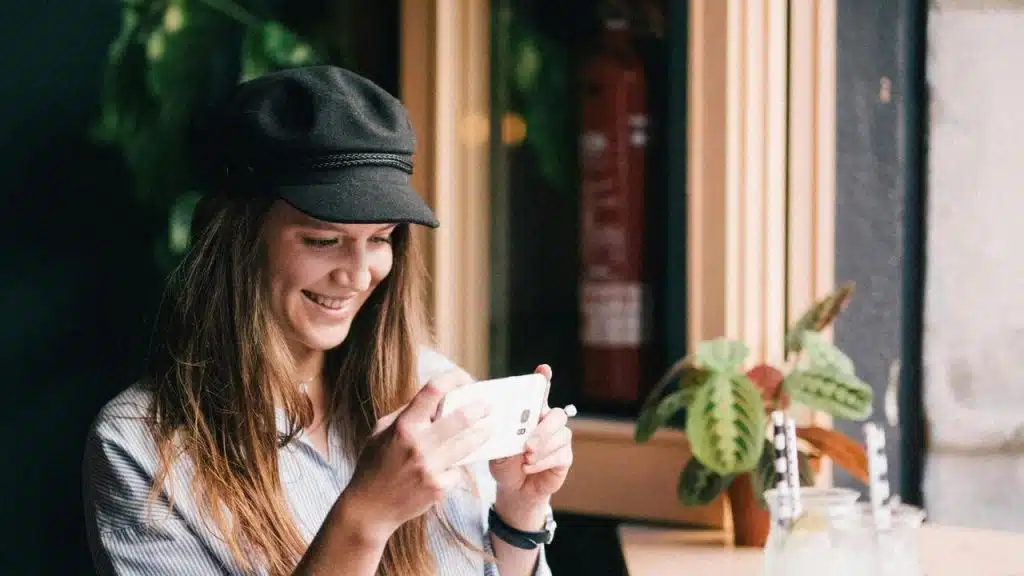 A smiling woman in a black cap looking at her phone by a café window.