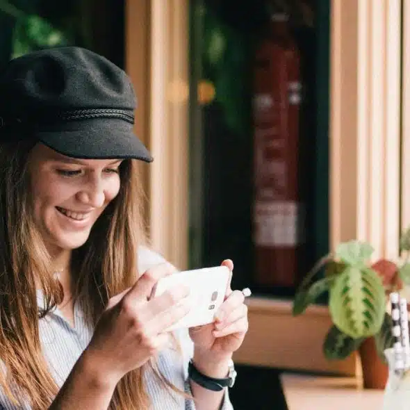 A smiling woman in a black cap looking at her phone by a café window.