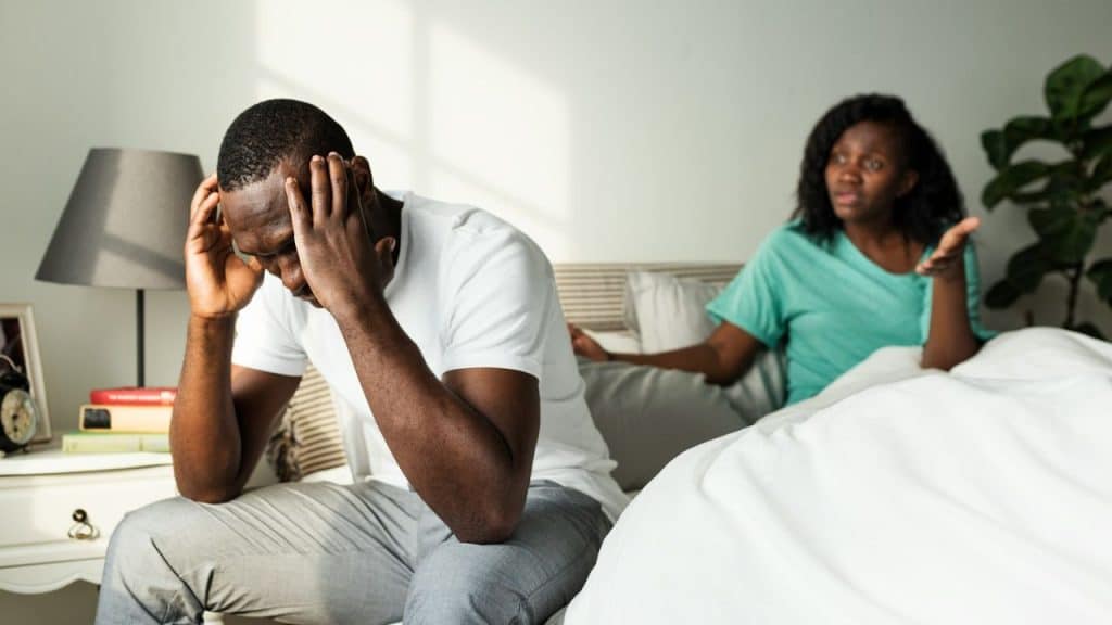 A man sits on a bed holding his head while a woman gestures nearby.