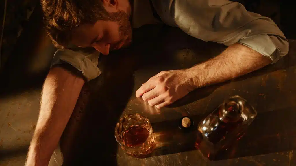 A man slumped over a table beside a glass and bottle of liquor.