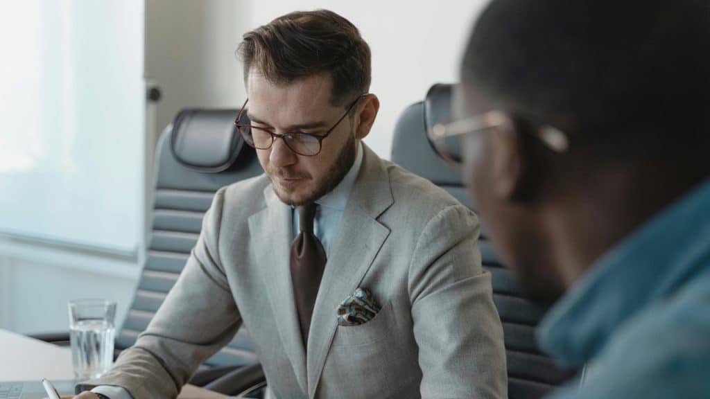 A man in a suit and glasses looks down while sitting at a conference table.