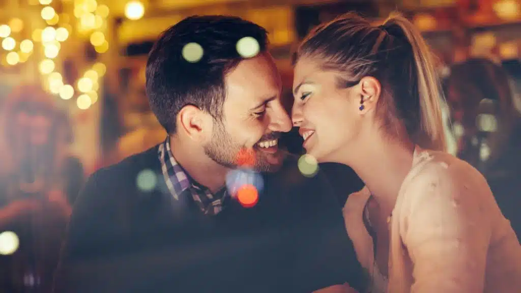 A man and woman smiling closely at each other in a dimly lit, festive setting.
