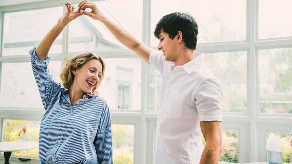 A man and woman hold hands and dance together in front of large windows.