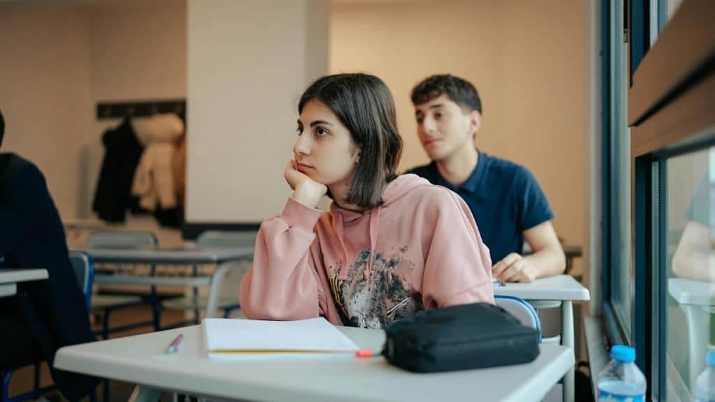 A young woman sitting at a desk in a classroom, resting her chin on her hand.