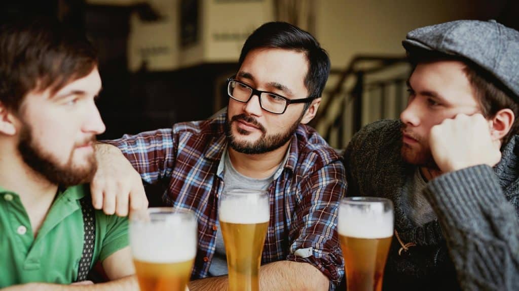 Three men sit at a table with three glasses of beer, engaged in a conversation.