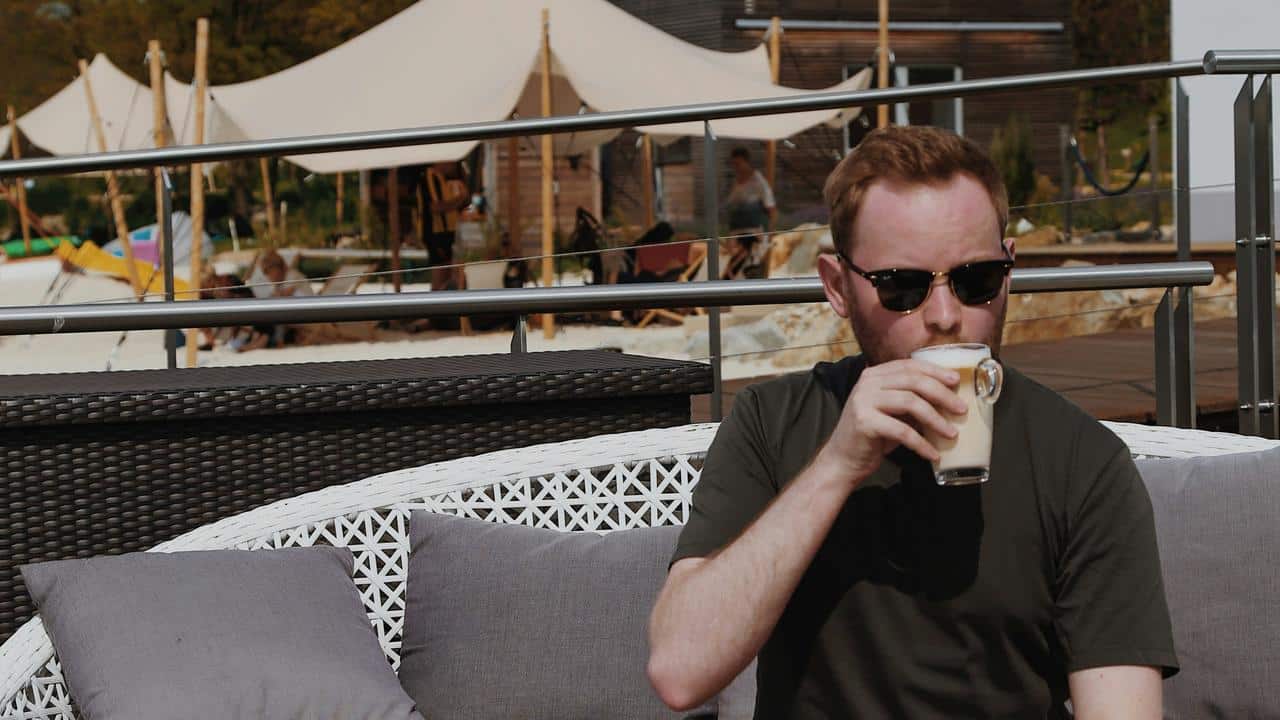 A man wearing sunglasses drinking coffee while sitting outdoors.