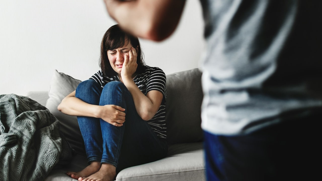 A woman is crying while sitting on a couch while her husband is standing in front of her.