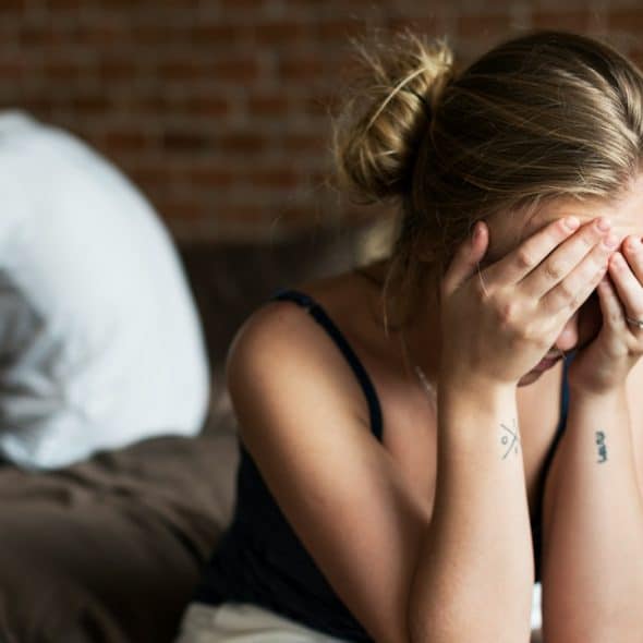 A distressed woman sits on a bed covering her face with her hands while a man sits turned away in the background, suggesting tension or conflict in a relationship.