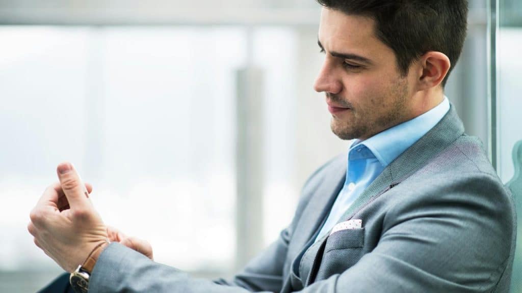A man in a suit adjusting his cuff while sitting indoors.