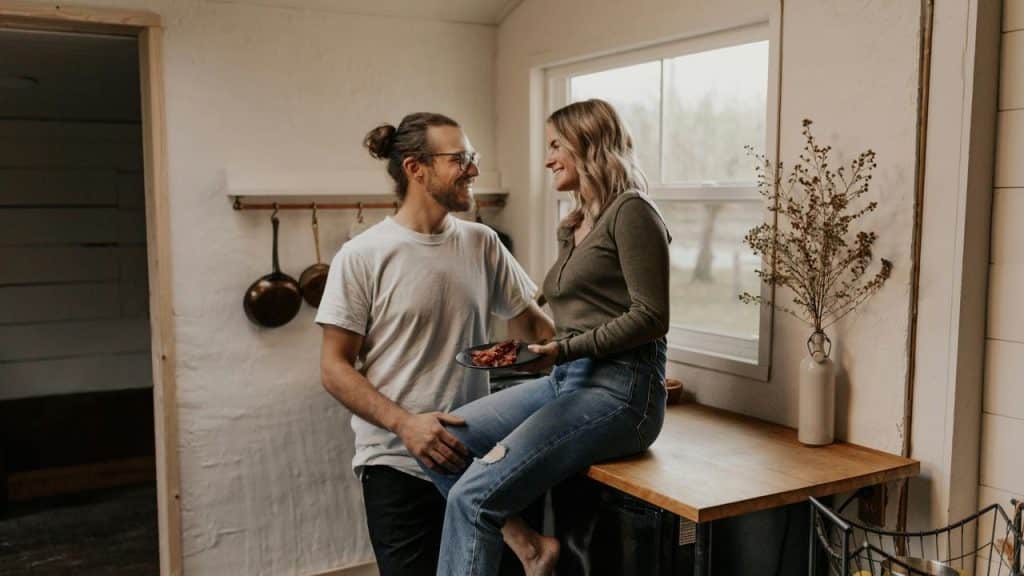 A couple smiling at each other while sharing food in a cozy kitchen.