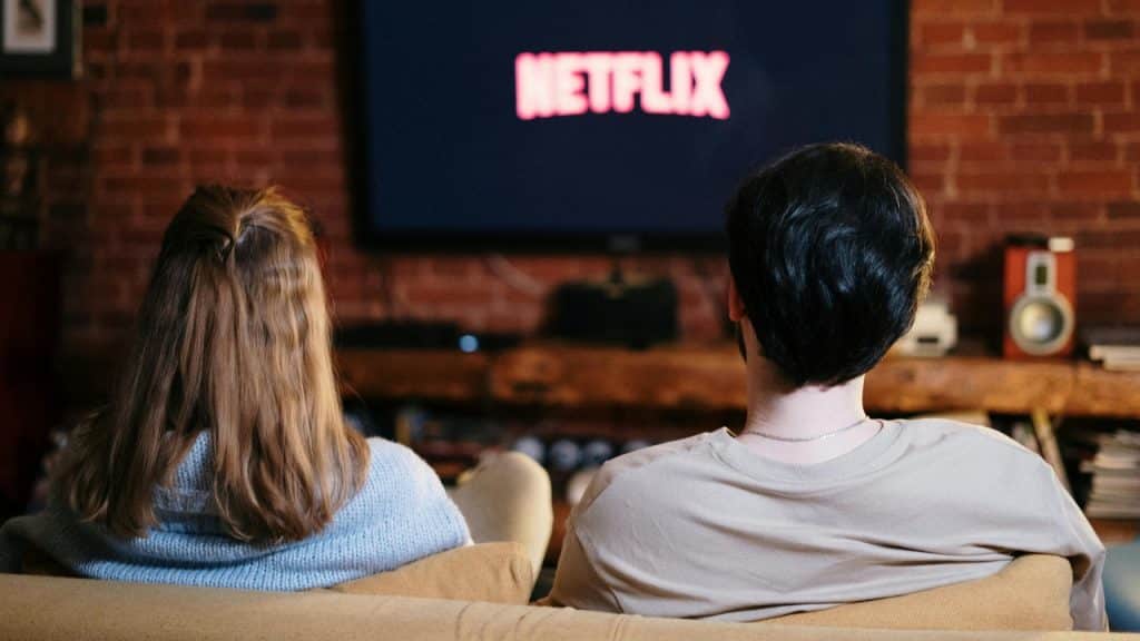 A couple sits on a couch from behind, watching a screen displaying the Netflix logo.
