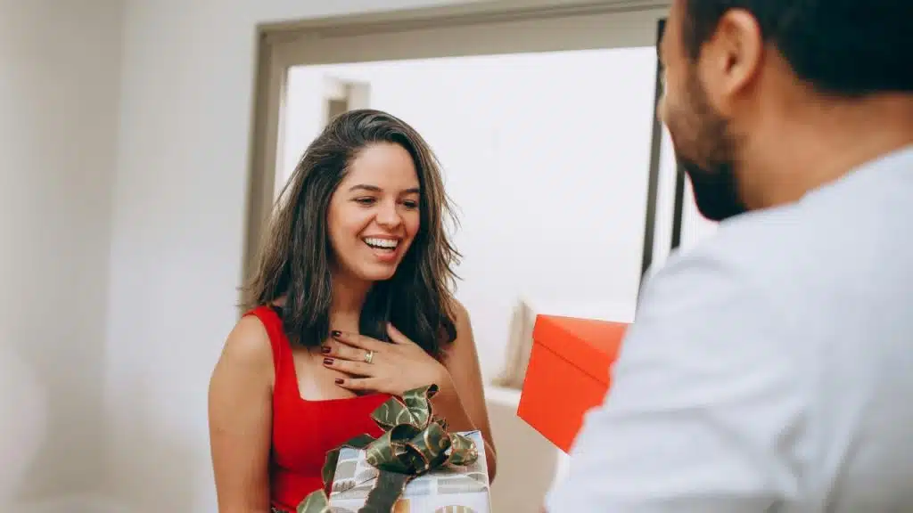 A smiling woman with a gift box holds her hand to her chest.