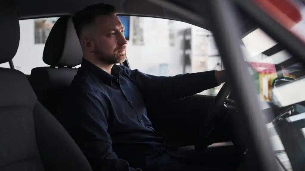 A bearded man in a dark shirt sits in a car looking out the window.