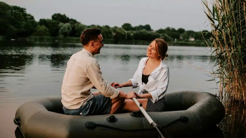 A couple sitting in a small boat on a calm lake, smiling and holding hands.