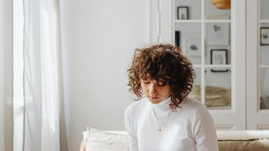 A person with curly hair sitting thoughtfully indoors.