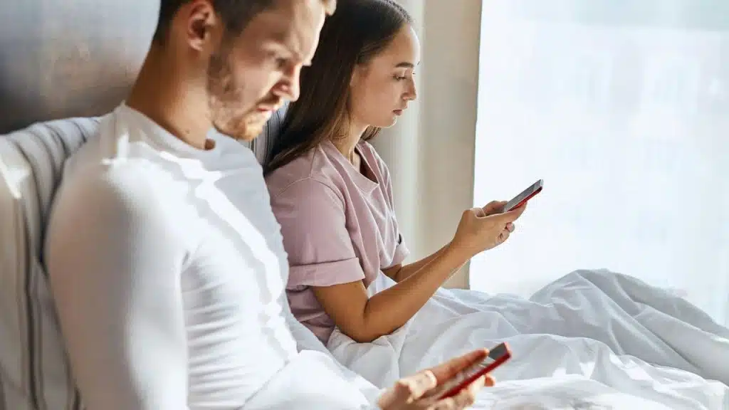 A man and woman sit in bed together, both focused on their individual mobile phones.