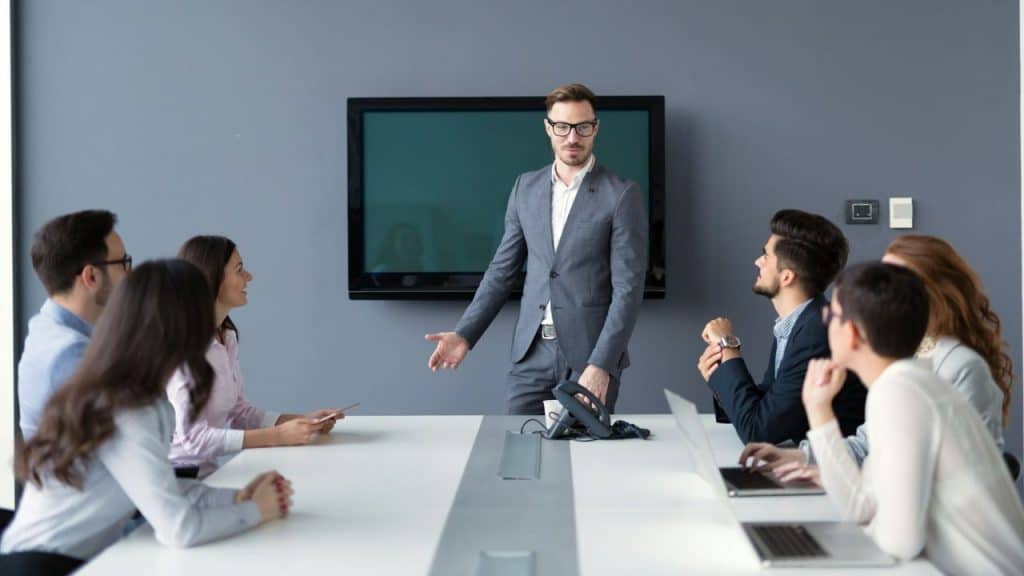 A man in a suit stands and gestures while leading a meeting with seated colleagues.