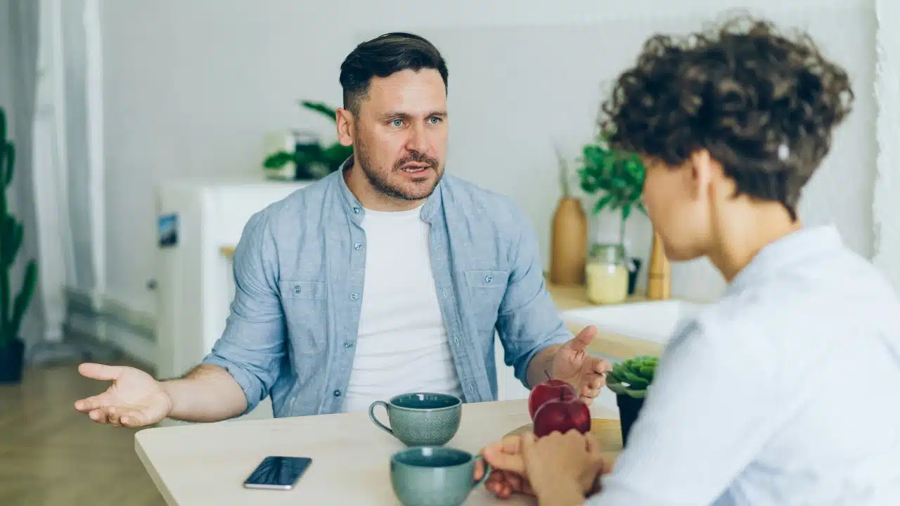 A man with a beard gestures with open hands while talking to a woman seated.