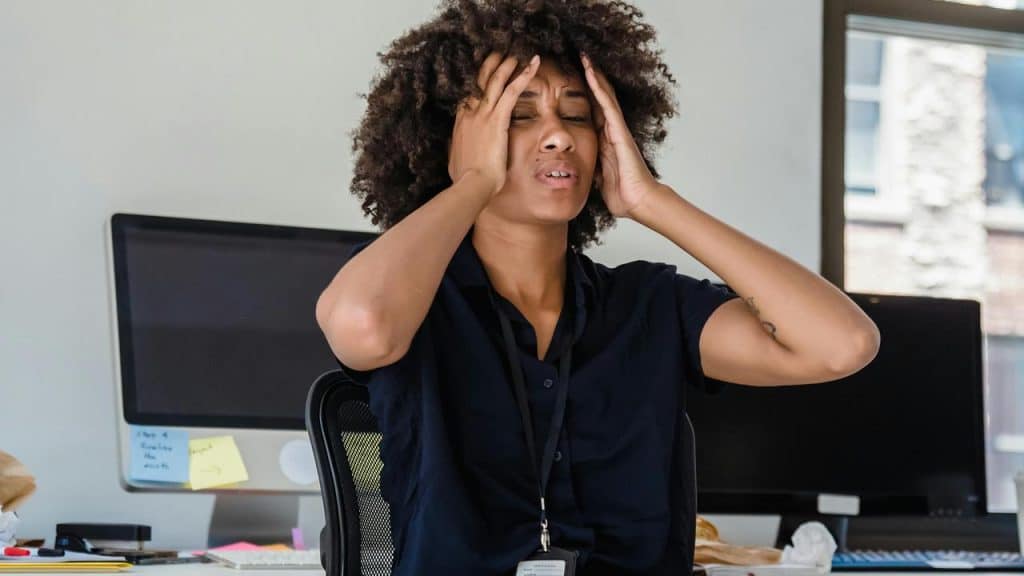 A stressed woman sits at a desk holding her head in an office.