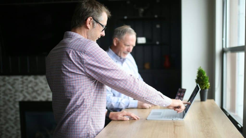 A man in a checkered shirt points at a laptop screen while another man looks on.