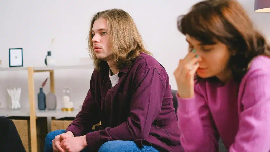 A man looks forward while a woman in the foreground rubs the bridge of her nose.